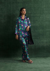 A woman standing against a green backdrop, wearing a navy blue silk pajama set with vibrant multicolor prints and pink flamingo designs. The outfit includes a button-down shirt and front pockets.